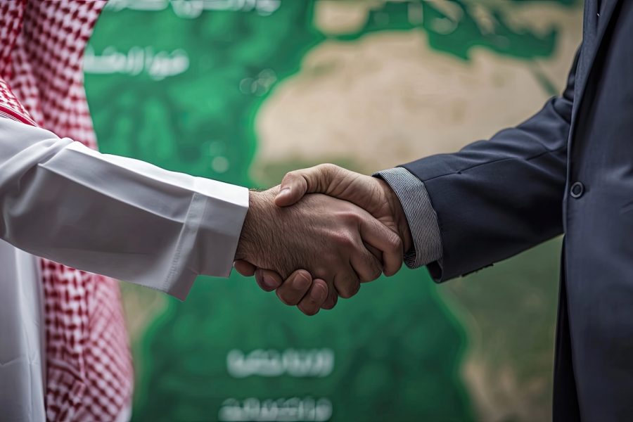 Handshake of a businessman in an official suit and a man in Saudi Arabian attire. No faces. In the background we see a green map of only Saudi Arabia